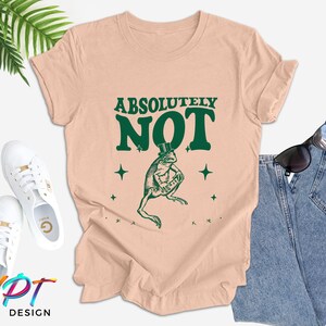 Puede incluir: Una camiseta rosa claro con una rana verde que lleva un sombrero de copa y el texto "Absolutamente no" en letras verdes.