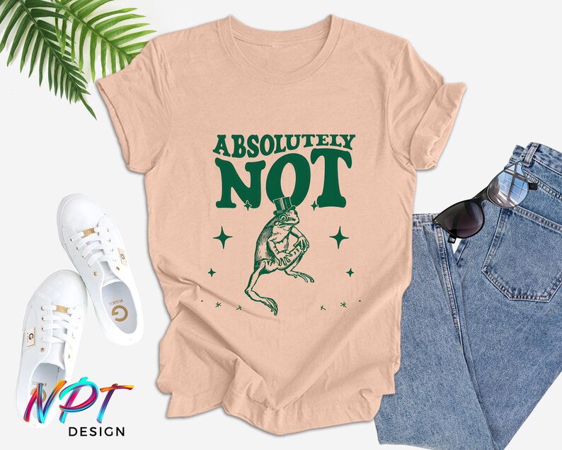 Puede incluir: Una camiseta rosa claro con una rana verde que lleva un sombrero de copa y el texto "Absolutamente no" en letras verdes.