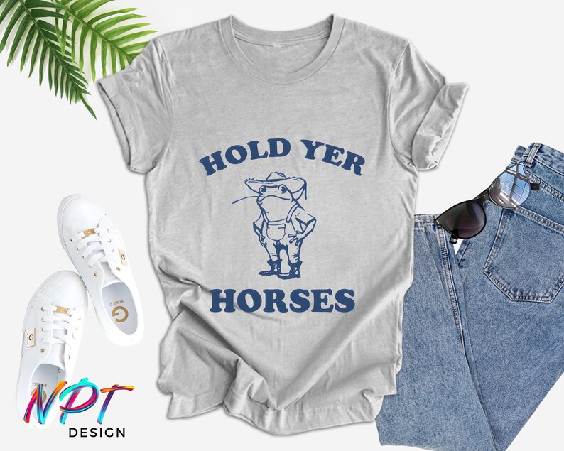 Puede incluir: Una camiseta gris claro con las palabras "HOLD YER HORSES" en azul marino, con una rana de dibujos animados con un sombrero de vaquero. La camiseta se combina con vaqueros azules, zapatillas blancas y gafas de sol.