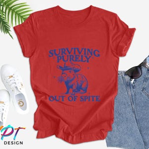 May include: Red t-shirt with a blue graphic of a cartoon animal wearing a sombrero and the text "Surviving Purely Out of Spite".
