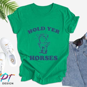 Puede incluir: Una camiseta verde con la frase azul "HOLD YER HORSES" y un dibujo de un vaquero. La camiseta se combina con vaqueros azules, zapatillas blancas y gafas de sol. El dise&ntilde;o es de NPT DESIGN.