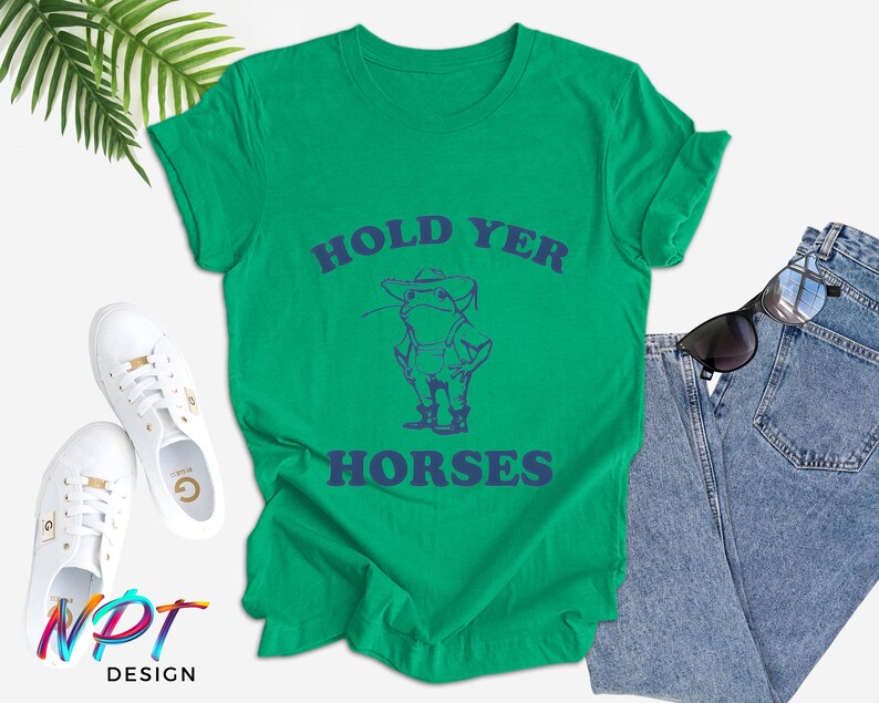 Puede incluir: Una camiseta verde con la frase azul "HOLD YER HORSES" y un dibujo de un vaquero. La camiseta se combina con vaqueros azules, zapatillas blancas y gafas de sol. El dise&ntilde;o es de NPT DESIGN.