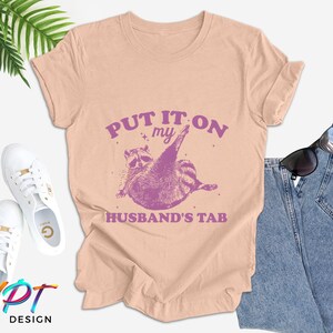 May include: A light pink t-shirt with the text "Put it on my husband's tab" in purple. A cartoon raccoon wearing a purple hat is shown above the text.
