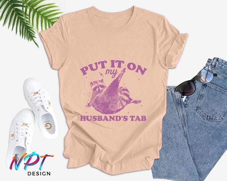 May include: A light pink t-shirt with the text "Put it on my husband's tab" in purple. A cartoon raccoon wearing a purple hat is shown above the text.