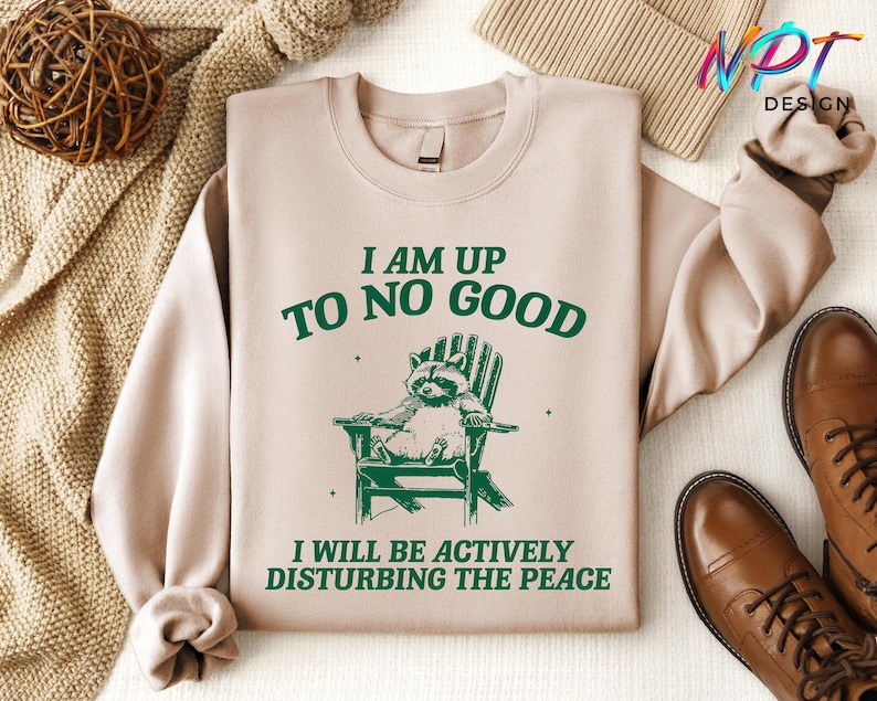 May include: A tan sweatshirt with a green graphic of a raccoon sitting in a chair. The text "I am up to no good. I will be actively disturbing the peace" is printed on the sweatshirt.