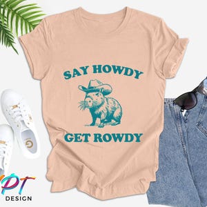 K&ouml;nnte beinhalten: Ein pfirsichfarbenes T-Shirt mit einem Aufdruck eines Capybaras, das einen Cowboyhut tr&auml;gt. Der Text "Say Howdy Get Rowdy" ist in T&uuml;rkis auf dem Shirt gedruckt.