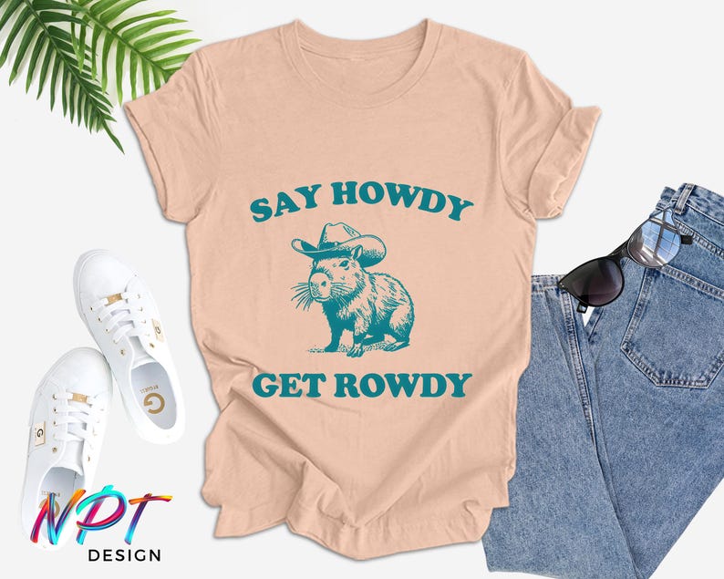 K&ouml;nnte beinhalten: Ein pfirsichfarbenes T-Shirt mit einem Aufdruck eines Capybaras, das einen Cowboyhut tr&auml;gt. Der Text "Say Howdy Get Rowdy" ist in T&uuml;rkis auf dem Shirt gedruckt.