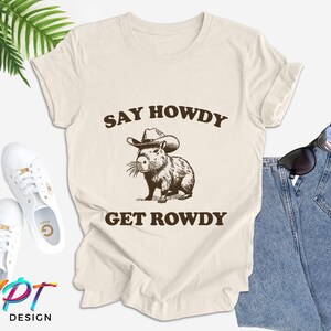 May include: A beige t-shirt with the text "Say Howdy Get Rowdy" printed on it. A cartoon capybara wearing a cowboy hat is in the center of the text.