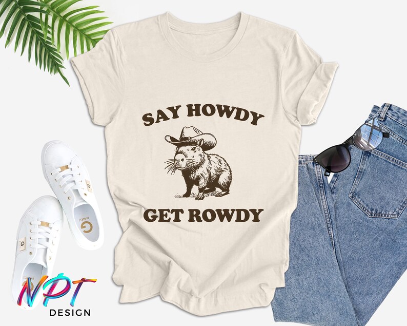 May include: A beige t-shirt with the text "Say Howdy Get Rowdy" printed on it. A cartoon capybara wearing a cowboy hat is in the center of the text.