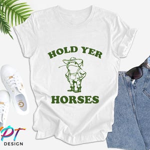 May include: White t-shirt with green text that says "Hold Yer Horses" and a cartoon frog wearing a hat and overalls.