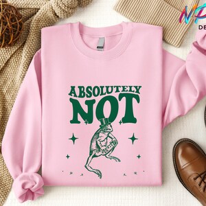 Puede incluir: Una sudadera rosa con el texto "ABSOLUTELY NOT" en letras verdes. Debajo del texto hay una rana verde con un sombrero de copa. La rana est&aacute; rodeada de peque&ntilde;as estrellas blancas.