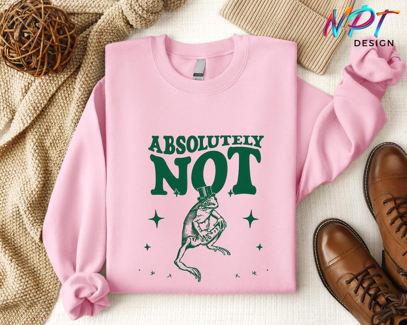 Puede incluir: Una sudadera rosa con el texto "ABSOLUTELY NOT" en letras verdes. Debajo del texto hay una rana verde con un sombrero de copa. La rana est&aacute; rodeada de peque&ntilde;as estrellas blancas.