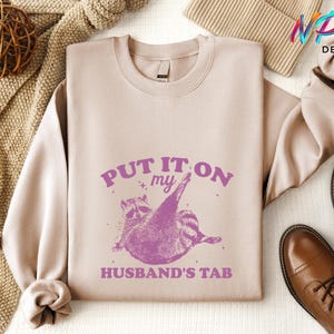 May include: A light pink sweatshirt with the text "PUT IT ON MY HUSBAND'S TAB" in purple lettering. A cartoon raccoon with a pink outline is holding a credit card in its paw.