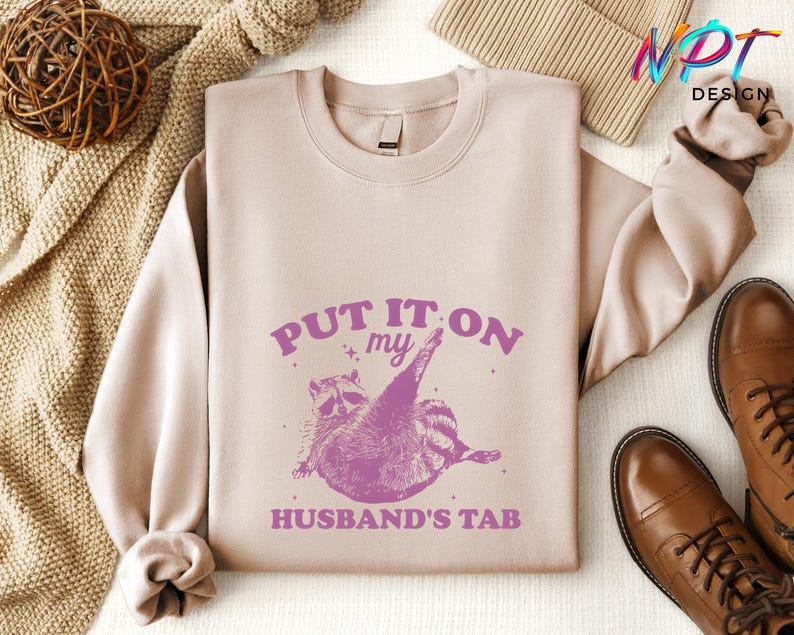 May include: A light pink sweatshirt with the text "PUT IT ON MY HUSBAND'S TAB" in purple lettering. A cartoon raccoon with a pink outline is holding a credit card in its paw.