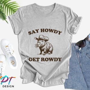 May include: A gray t-shirt with the text "Say Howdy Get Rowdy" and a cartoon image of a capybara wearing a cowboy hat.