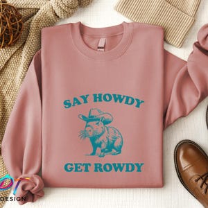 K&ouml;nnte beinhalten: Ein rosa Sweatshirt mit dem Text "Say Howdy Get Rowdy" in blaugr&uuml;nen Buchstaben. Ein Cartoon-Capybara mit Cowboyhut befindet sich in der Mitte des Textes.
