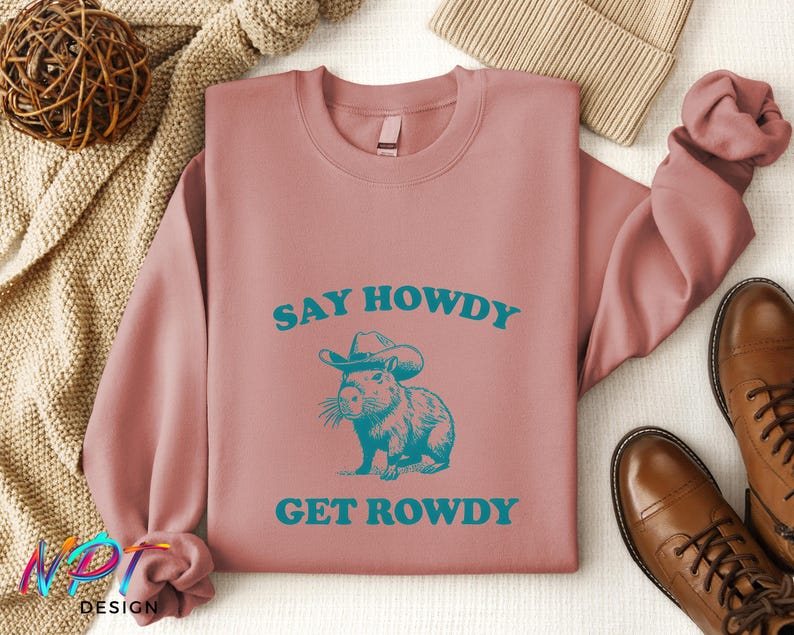 K&ouml;nnte beinhalten: Ein rosa Sweatshirt mit dem Text "Say Howdy Get Rowdy" in blaugr&uuml;nen Buchstaben. Ein Cartoon-Capybara mit Cowboyhut befindet sich in der Mitte des Textes.