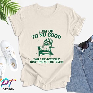 May include: A beige t-shirt with a green graphic of a raccoon sitting in a deckchair. The text "I am up to no good" and "I will be actively disturbing the peace" is printed on the shirt.