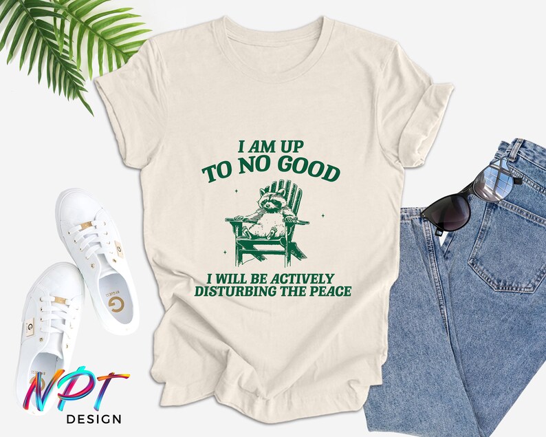 May include: A beige t-shirt with a green graphic of a raccoon sitting in a deckchair. The text "I am up to no good" and "I will be actively disturbing the peace" is printed on the shirt.
