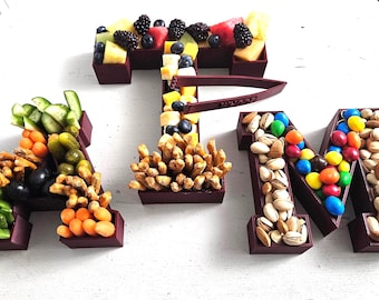 Op de afbeelding: Een creatieve opstelling van voedselitems die het woord "ATM" spellen. De letter "A" is gevuld met groenten en snacks, "T" met fruit en "M" met noten en snoep. De letters staan in een donkerbruine bak.