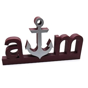 May include: A maroon-colored decorative sign with the letters "a m" and a silver anchor in the center. The anchor is positioned between the letters, adding a nautical theme to the sign.