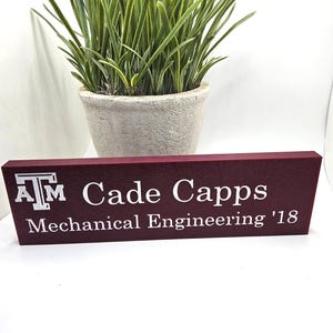 May include: A maroon desk sign with white text that reads "ATM Cade Capps Mechanical Engineering '18". A potted plant with green grass-like leaves sits behind the sign in a light-colored pot.
