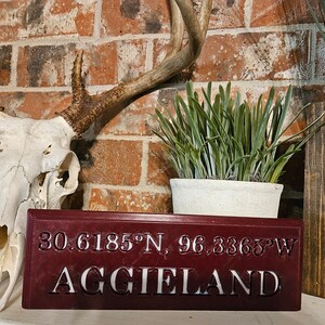 May include: A maroon wooden sign with white lettering that reads "30.6185°N, 96.3365°W AGGIELAND". The sign is sitting on a table in front of a brick wall and a deer skull.