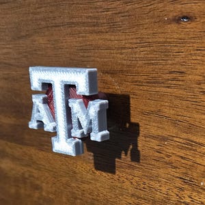 May include: A white 3D printed "TAM" logo with a red background. The logo is attached to a wooden surface.
