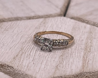 May include: A gold ring featuring a square-cut diamond centre stone and smaller diamonds on the band. The ring is set on a light-coloured, textured surface. The ring is marked "K 18K".