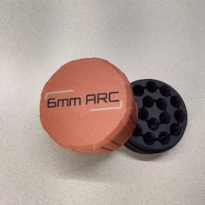 May include: A copper-colored, circular object with the text "6mm ARC" in white. Next to it is a black, circular object with multiple holes. The objects are on a light gray surface.