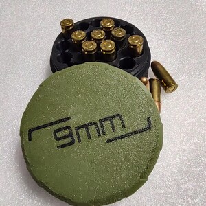 May include: A black 9mm ammunition holder with a green lid. The lid has the text "9mm" printed in black. The holder contains several brass-colored bullets. Some bullets are outside the holder.