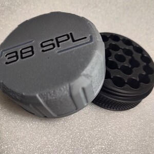 May include: A gray, cylindrical object with the text "38 SPL" printed on the top. Next to it is a black, circular object with multiple holes. The objects are likely related to ammunition.