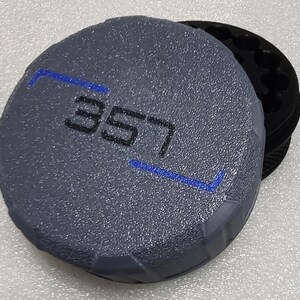 May include: A gray, textured, round object with the number "357" in black and blue accents. The object is partially covering a black, circular base with multiple holes. The design has a geometric pattern.