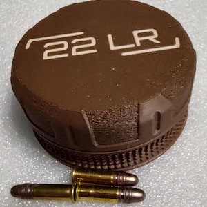 May include: A brown, cylindrical object with the text "22 LR" in white, alongside two .22 LR bullets. The object appears to be a lid or cap, possibly for ammunition storage or a related item. The bullets are gold-colored with a silver tip.