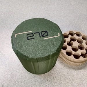 May include: A green cylindrical container with the number "270" printed on the top, next to a beige circular tray with multiple holes. The container has a textured surface. The tray has a ribbed edge and is open, revealing the interior.