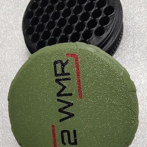 May include: A black, circular container with multiple holes sits above a green, circular object. The green object has the text "22 WMR" in black with red accents. The items appear to be made of plastic.
