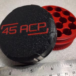 May include: A black, textured object with the red text "45 ACP" is next to a red, circular object with multiple holes. The objects are on a light surface, with a ruler in the foreground.
