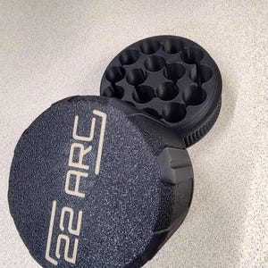 May include: A black, textured container with the text "22 ARC" in white. The container is open, revealing a series of circular compartments. The object is on a light gray surface.