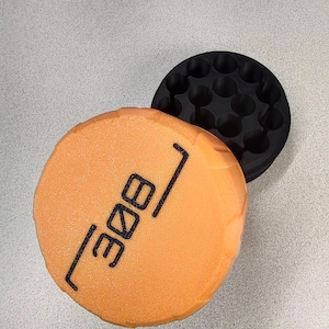 May include: An orange, circular object with the number "308" printed in black, next to a black, circular object with multiple holes. The objects are on a light gray surface.