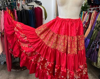 May include: A vibrant red tiered skirt with gold floral accents and ruffled edges. The skirt is displayed on a mannequin, showcasing its flowing design. The skirt's fabric has a shiny, satin-like appearance, suggesting a festive or bohemian style.