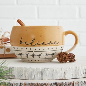 May include: A large, tan ceramic mug with the word "believe" in black script. The bottom half of the mug is white with a black star and dot pattern. A cinnamon stick rests inside the mug. Two small pinecones sit near the mug.