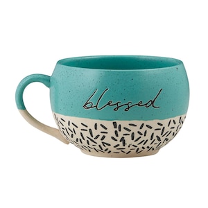 May include: A teal ceramic mug with the word "blessed" in cursive script. The top half of the mug is teal with a speckled texture, while the bottom half is cream-colored with black dashes. The handle is cream-colored.
