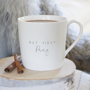 May include: White ceramic mug with the text "BUT FIRST Pray" in black script. The mug is filled with a dark liquid and sits on a wooden slice with two cinnamon sticks. The mug has a curved handle.