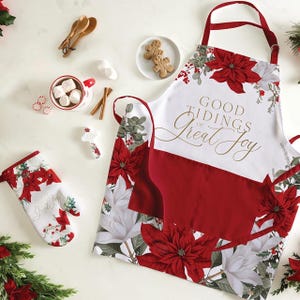 May include: A festive Christmas-themed kitchen set featuring a white apron and oven mitt with red poinsettias and the text "Good Tidings of Great Joy." The set includes a red pocket, and is accompanied by gingerbread cookies, cinnamon sticks, and a mug of marshmallows.