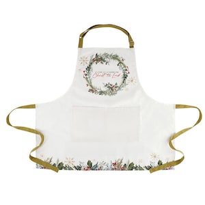 May include: White apron with a Christmas wreath design and the text "O Come Let Us Adore Him Christ the Lord." The apron has two pockets and olive green straps and trim. The bottom of the apron has a floral and berry border.