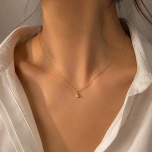 May include: A delicate gold chain necklace featuring a single, small, round, clear gemstone pendant centered at the base of the neck. The necklace is worn against a white collared shirt, highlighting the thin metallic links and the sparkling stone.