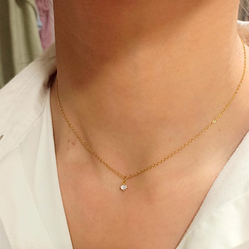 May include: A delicate gold chain necklace featuring a single, small, round, clear gemstone pendant centered on the chain. The necklace rests against the neck, showcasing a thin, polished metal link design with a secure clasp connection point.