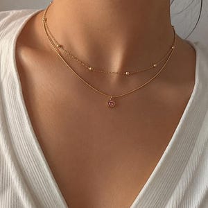 Könnte beinhalten: Eine goldfarbene, mehrlagige Halskette bestehend aus zwei verschiedenen Ketten. Die obere Kette ist mit kleinen, gleichmäßig verteilten Goldperlen verziert, während die untere Kette einen runden Anhänger mit einem kleinen, rosa Edelstein trägt.
