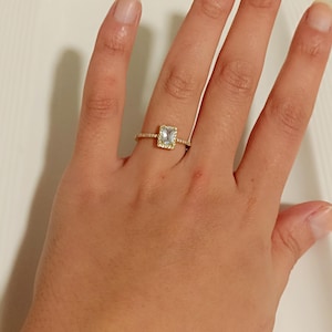 Könnte beinhalten: Ein goldener Ring mit einem rechteckigen, hellblauen Edelstein, umgeben von kleineren Steinen. Der Ring befindet sich an einer Hand, wobei das Design und die Details des Schmucks hervorgehoben werden.