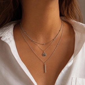 Könnte beinhalten: Eine mehrlagige silberfarbene Halskette bestehend aus drei verschiedenen Ketten. Die oberste Kette hat kleine Metallperlen, die mittlere Kette trägt einen runden Anhänger und die unterste Kette einen schmalen, vertikalen Stabanhänger.